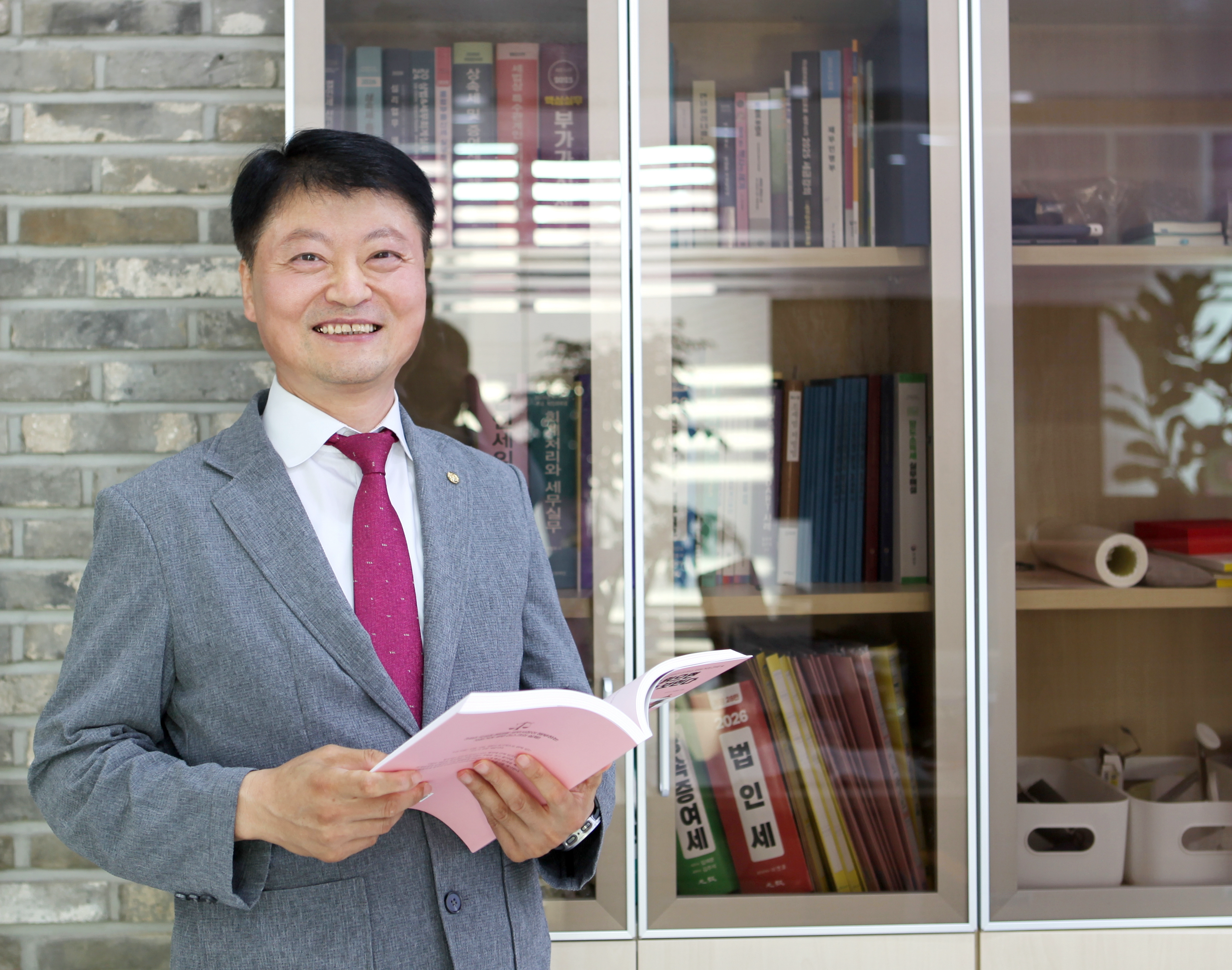 사무실 책장 앞에서 책을 펼쳐 들고 웃고 있는 우창용 세무사